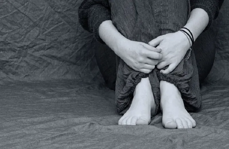 Black and white photo of a barefoot person sitting on the floor, hugging their knees